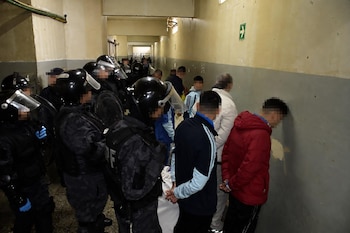 Agentes con equipo antidisturbios vigilan a varios hombres alineados frente a la pared con las manos a la espalda en un pasillo