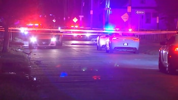 Varios coches de policía con luces rojas y azules encienden la noche en una calle acordonada con cinta amarilla. Se observan señales de tráfico y edificios