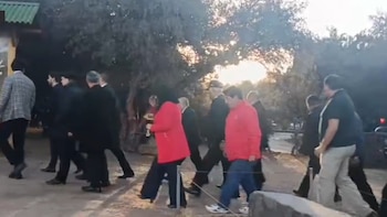 Grupo de personas camina en un área al aire libre con árboles y vegetación. Algunas personas visten trajes oscuros y otras ropa casual