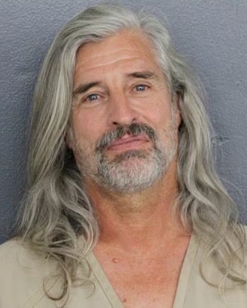 Primer plano de un hombre con cabello gris largo y barba. Viste una camisa de color claro. El fondo es de color gris