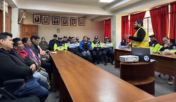Un hombre con chaleco amarillo habla ante un grupo de personas sentadas alrededor de mesas de madera en una sala con cortinas rojas