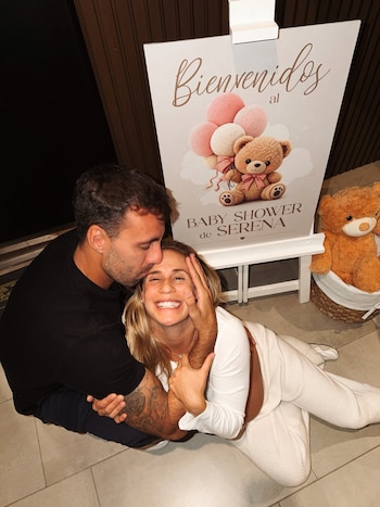 Un hombre besa la frente de una mujer rubia sonriente en un baby shower. Detrás, un cartel de 'Bienvenidos al Baby Shower de Serena' y un oso de peluche