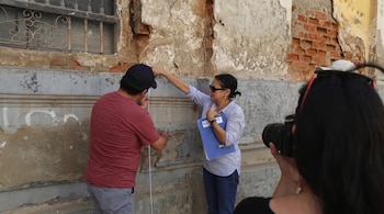 Un hombre y una mujer con una cinta métrica y una carpeta examinan una pared con ladrillos expuestos mientras los fotografían.