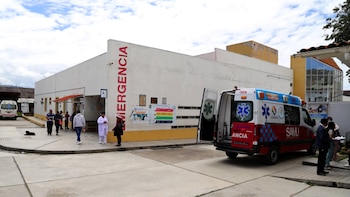 Exterior del Hospital Simón Bolívar con una ambulancia blanca y roja estacionada, sus puertas traseras abiertas, y personas caminando cerca de la entrada de Emergencia