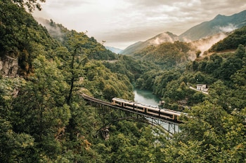 Un tren blanco y amarillo cruza un puente de acero sobre un río en un valle montañoso cubierto de densa vegetación verde, con nubes bajas en las cimas