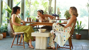 Tres mujeres sentadas en una mesa de madera en una habitación luminosa con grandes ventanas y plantas. Visten ropa casual y sonríen mientras interactúan