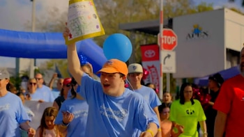 Joven sonriente con gorra naranja y camiseta azul levanta un cartel en evento, rodeado de personas, un arco inflable azul y un globo