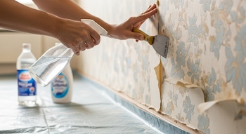 Dos manos, una rocía una pared con papel tapiz floral y la otra usa una espátula para despegarlo. Hay un plástico protector en el suelo y dos botellas en el fondo.