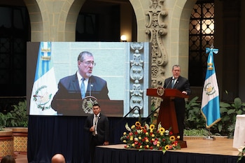 Un hombre de traje habla en un atril, una pantalla grande muestra su imagen, un guardia de seguridad, una bandera de Guatemala y arreglos florales