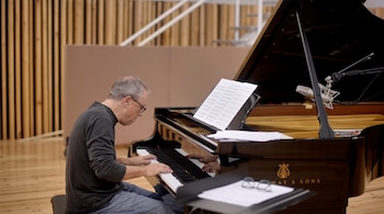 Un hombre con gafas toca un piano de cola negro con partituras abiertas y micrófonos en un estudio con paredes de madera
