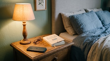 Mesa de madera con una lámpara encendida, anteojos, un libro y un teléfono boca abajo, junto a una cama con almohadas.