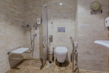 Baño con paredes de mármol beige, inodoro, lavabo, asiento de ducha plegable, ducha con manguera, barras de sujeción metálicas y teléfono