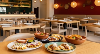 Interior de restaurante con mesas de madera, sillas, bar al fondo. Primer plano de mesa con un plato de chilaquiles, un cuenco de pozole, tacos y sopes.