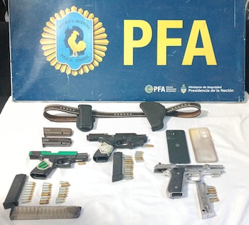 Vista cenital de tres armas de fuego, cargadores, municiones, un cinturón con funda y dos teléfonos celulares, frente a un cartel de la PFA