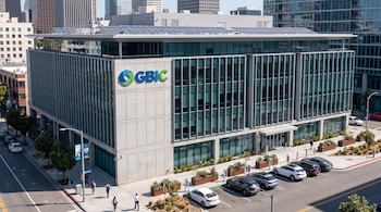 Edificio moderno de GBIC con paneles solares en el techo, rodeado de aceras, personas, autos estacionados con cargadores eléctricos y edificios urbanos.
