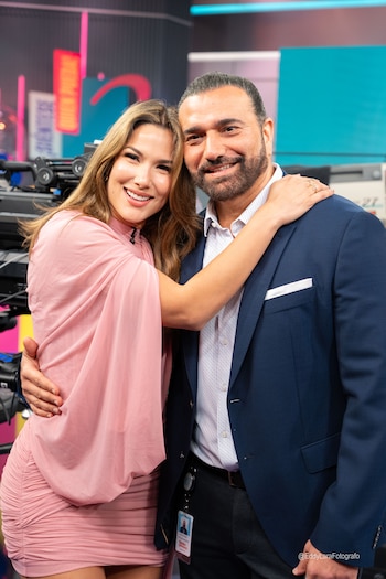 Alejandra Jaramillo, con vestido rosa, abraza a Carlos Mesber, quien viste traje azul, en un estudio de televisión con luces de colores