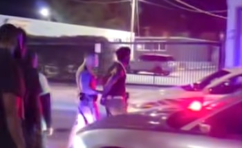 Un agente de policía camina junto a un hombre esposado cerca de patrullas con luces rojas encendidas por la noche