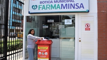 Mujer con blusa gris y pantalón azul deposita una caja en un contenedor rojo de medicamentos frente a una farmacia "Farma Minsa"
