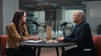 Demis Hassabis, un hombre calvo con gafas, y una mujer sentados en una mesa con dos micrófonos, conversando con un juego de Jenga entre ellos