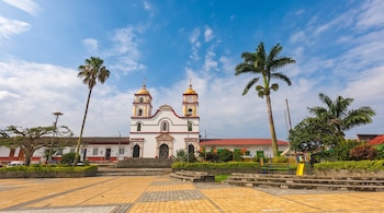 Iglesia de fachada blanca con cúpulas amarillas, dos palmeras altas, una plaza pavimentada con baldosas amarillas y cielo azul.