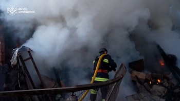 Un bombero con uniforme oscuro y franjas reflectantes, con una manguera amarilla, trabaja entre columnas de humo blanco y gris de una estructura dañada con escombros y fuego