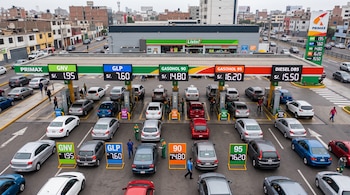 Vista aérea de una gasolinera Primax con carteles de precios de combustibles y varias filas de automóviles junto a los surtidores.
