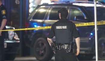 Un oficial de policía de Yonkers de espaldas, con uniforme negro y gorra, parado frente a un vehículo policial y cinta amarilla que dice 'POLICE LINE DO NOT CROSS'