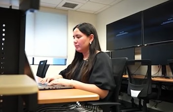 Una mujer sentada en un escritorio escribe en una computadora portátil dentro de un aula con monitores en la pared