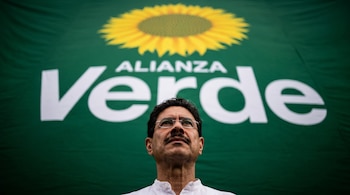 Iván Cepeda, de cerca y en contrapicada, con gafas y camisa blanca, tiene detrás un enorme cartel verde del partido Alianza Verde con un girasol amarillo.