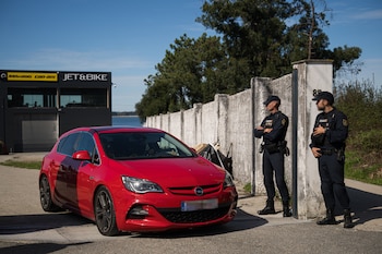 Dos agentes de la Policía Nacional uniformados de oscuro se encuentran junto a un coche rojo aparcado frente a un muro y un edificio, con un cielo azul de fondo