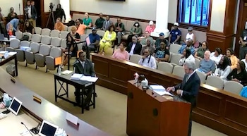 Un hombre en traje habla ante un podio de madera en una sala de audiencias frente a personas sentadas en el público