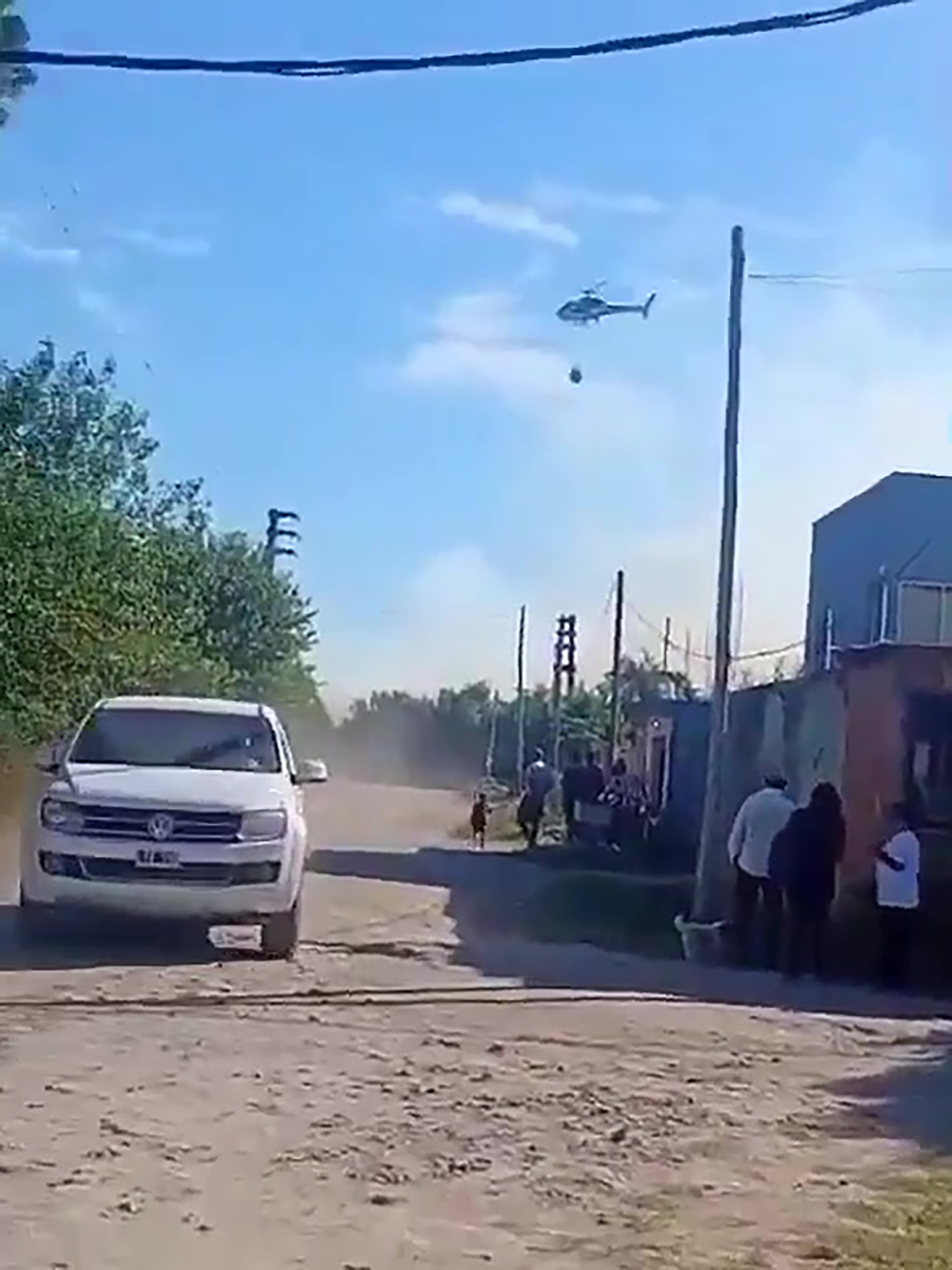 Un helicóptero hidrante opera sobre el Parque Pereyra Iraola, combatiendo los incendios forestales mientras una camioneta circula por una calle de tierra y vecinos observan la situación. (Captura de video)