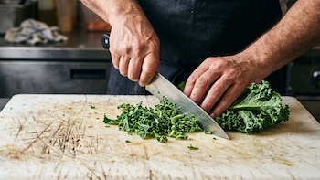 Manos de una persona que corta hortalizas de hoja verde con un cuchillo de metal sobre una tabla de picar blanca
