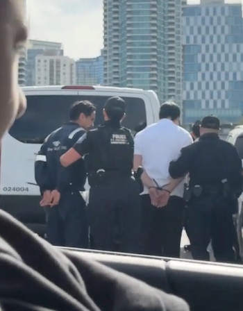 Vista de tres agentes de policía que detienen y esposan a un hombre vestido de blanco frente a un vehículo blanco, con edificios altos al fondo