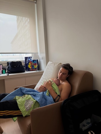Paulo Dybala, sin camisa, sentado en un sillón marrón claro, acuna a un bebé recién nacido envuelto en mantas verde y azul dentro de una habitación iluminada
