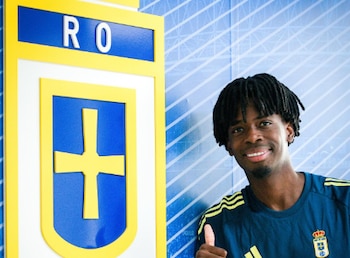 Hombre con peinado de rastas y camiseta deportiva azul sonríe y levanta el pulgar junto a una placa con el escudo del Real Oviedo