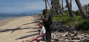 Un oficial uniformado interroga a un hombre sentado en una playa de arena. Detrás, hay palmeras, vegetación y el océano con barcos en la distancia. La playa tiene madera flotante