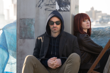 Charlie Cox con capucha y gafas de sol, y Deborah Ann Woll con chaqueta de cuero, sentados contra una pared de hormigón con grafitis y un fondo borroso