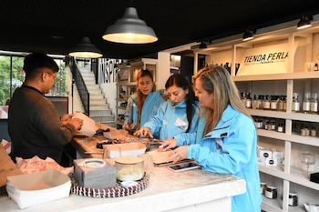 Cuatro personas, tres mujeres en chaquetas azules y un hombre, interactúan en el mostrador de un comercio. Estantes con productos visibles al fondo