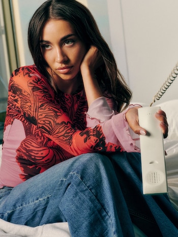 María Becerra sentada, vestida con una blusa de manga larga roja y negra con detalles rosas y jeans azules, sosteniendo un teléfono fijo blanco