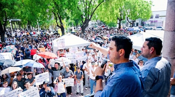 Gran multitud de personas reunidas al aire libre, con árboles, paraguas, pancartas y dos hombres en primer plano, uno con micrófono y brazo levantado.