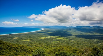 Vista aérea de una extensa costa con playa, océano azul, áreas verdes de bosques y praderas, montañas en el horizonte y un cielo con nubes.