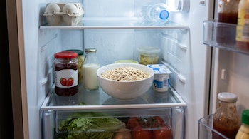 Un tazón de avena, un frasco de mermelada de fresa y botellas sobre la repisa de un refrigerador con tomates y lechuga en el cajón.