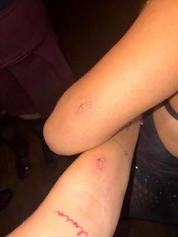 Dos brazos muestran pequeños tatuajes idénticos de mariposa en tinta negra. Uno de los brazos tiene un segundo tatuaje en rojo con una palabra