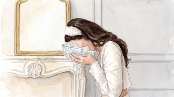Mujer de cabello largo con una diadema blanca, sumerge su rostro en un cuenco transparente con cubos de hielo y agua, frente a un espejo dorado y un mueble blanco.