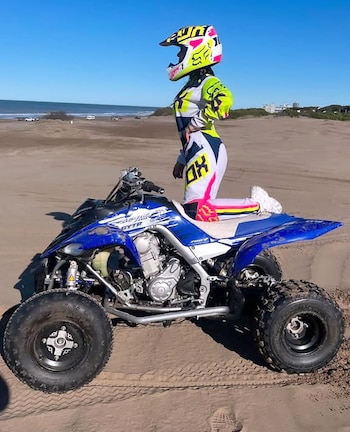 Una persona con equipo de motocross brillante y un casco se arrodilla sobre un cuatriciclo azul en una playa arenosa bajo un cielo despejado