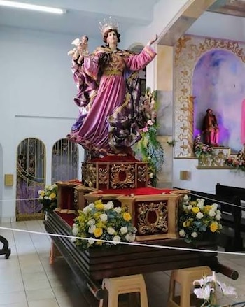 Estatua de una mujer con corona y un bebé en brazos, vestida con túnica morada y dorada, sobre una plataforma con flores blancas y amarillas en un interior