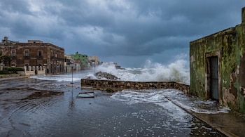 Olas grandes rompen contra el malecón de una ciudad costera. Hay edificios antiguos, charcos, una calle inundada, escombros y cielo gris con nubes de tormenta.