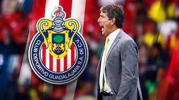 Guillermo Almada en traje gris da indicaciones junto al escudo del Club Deportivo El director técnico no ha tenido un saldo positivo enfrentando al rebaño. sobre un fondo de afición difuminado.