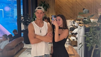 Brenda Di Aloy, con un vestido negro, sostiene un teléfono como si tomara una selfie junto a Sebastián Freidzon, con gorra y camiseta blanca, en una tienda de ropa